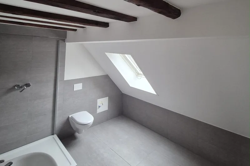 Badlüftung: Dachgeschossbad mit Dachfenster in der Schräge, Wand-WC und dunklen Deckenbalken